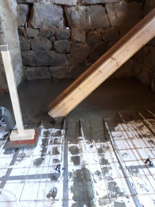 Nyutgjuten betong på isolerad golvyta i en byggarbetsplats med stenmur och träkonstruktion.