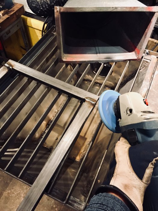 Person använder en vinkelslip på en metallstång över ett downdraft-bord i verkstad med synlig sugfläkt och slitage.