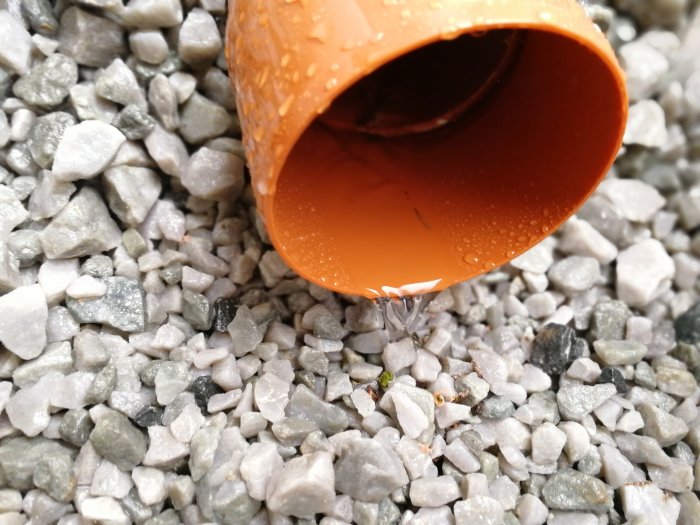 Slutet av ett orange dagvattenrör som ligger på en bädd av gråa singelstenar, med små vattendroppar synliga på röret.