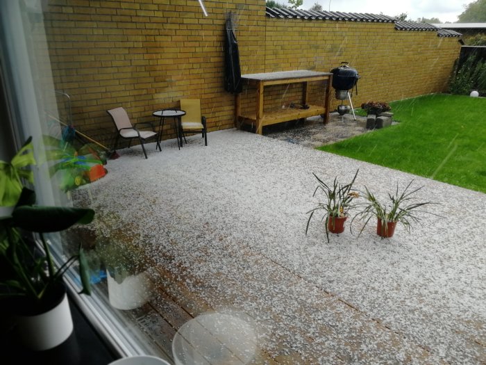 En bakgård täckt med hagel, med en grill, sittgrupp, och gula tegelväggar, sett från ett fönster.
