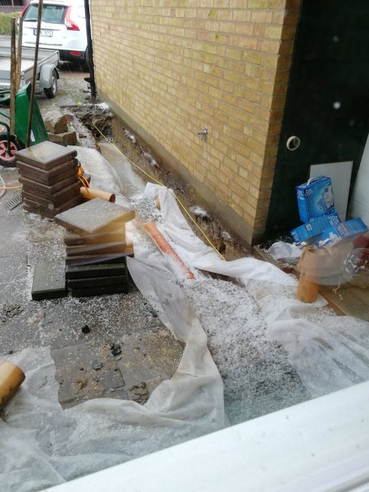 En byggarbetsplats vid en husvägg med ett täckt dike, byggmaterial och hagel på presenning.