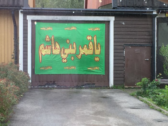 Grön skylt med arabisk text hängandes på husvägg, vilket tycks vara en religiös högtid.