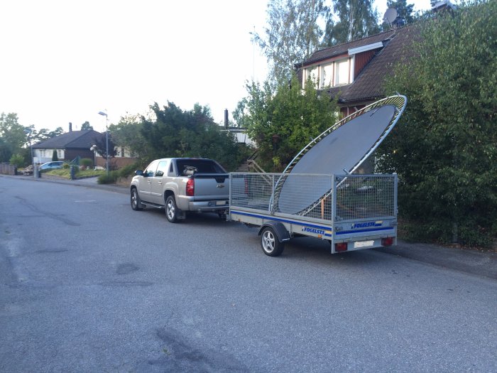 SUV med släpkärra lastad med stor satellitantenn på en bostadsgata.