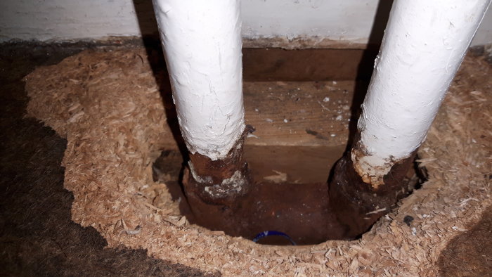 Rost på äldre värmeledningsrör som går genom ett hål i ett golv, omgivet av isoleringsmaterial.