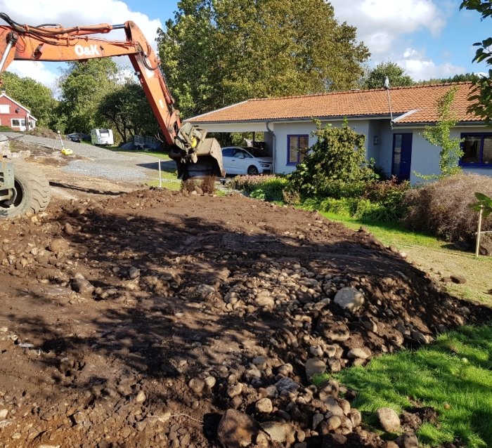 Grävmaskin som arbetar med schaktning av grusig mark nära hus och anlagd väg.