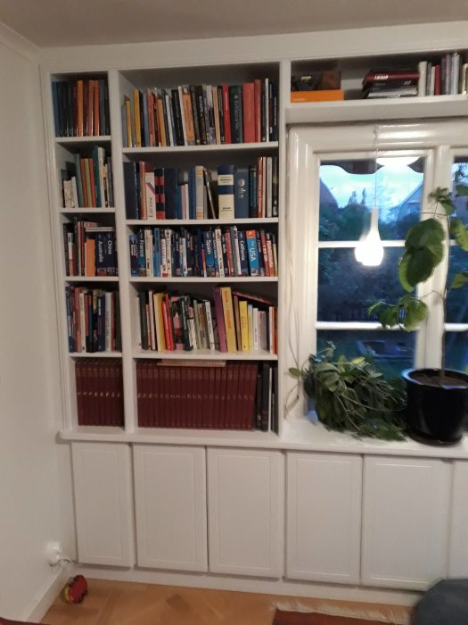 Vit bokhylla fylld med böcker bredvid ett fönster med växt på fönsterbrädan och skåp nedanför.