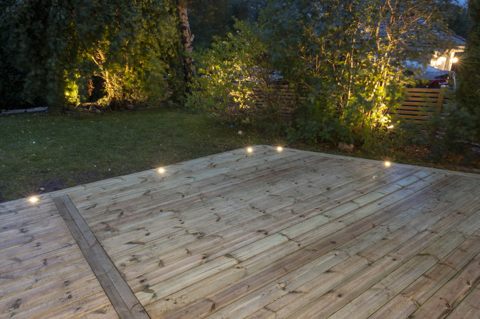 Ett nyligen renoverat träträdäck med infällda spotlights, omgivet av en välhållen trädgård i skymningen.