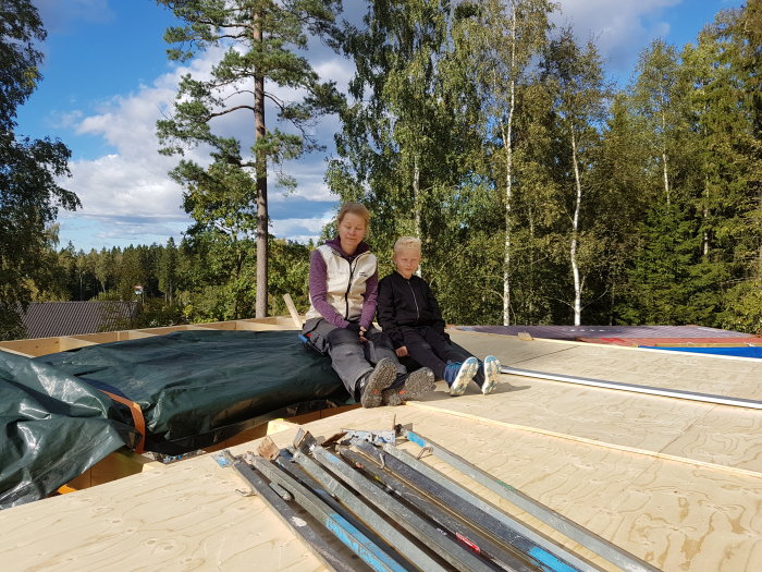 Två personer sitter på en halvfärdig balkongkonstruktion med skog i bakgrunden.