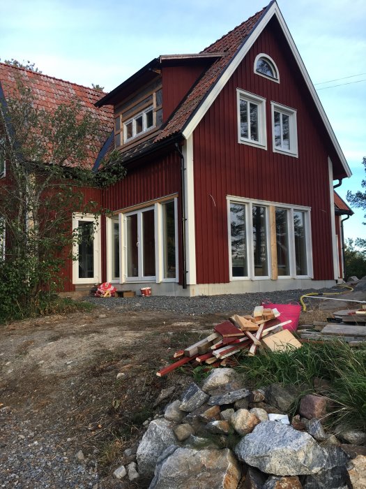 Rödmålat hus med lockläkt och hängrännor, byggmaterial på marken och takkupor synliga.