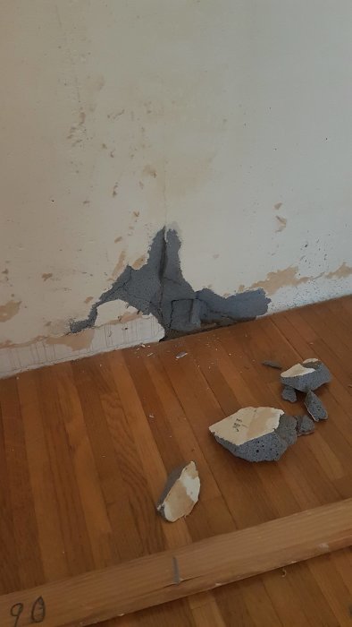 Skadad vägg med fallna putsbitar på trägolv i gammal lägenhet.