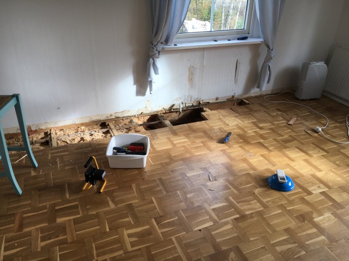 Uppbrutet trägolv med synliga skador och fukt, verktygslåda och avfuktare i ett rum i behov av renovering.