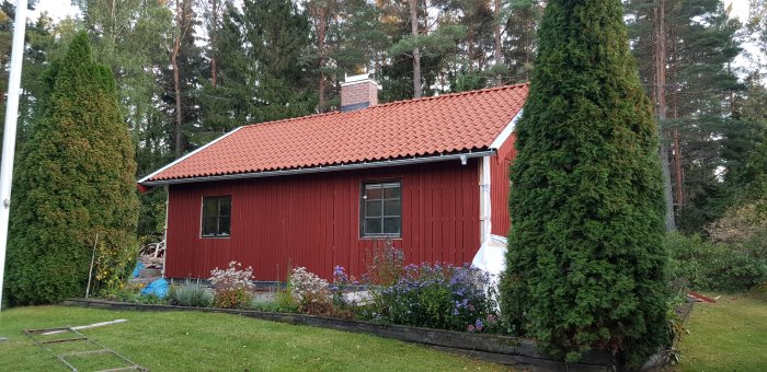 Renoverat rött trähus med nytt tegeltak, omgivet av grönska och blomster i en trädgård.