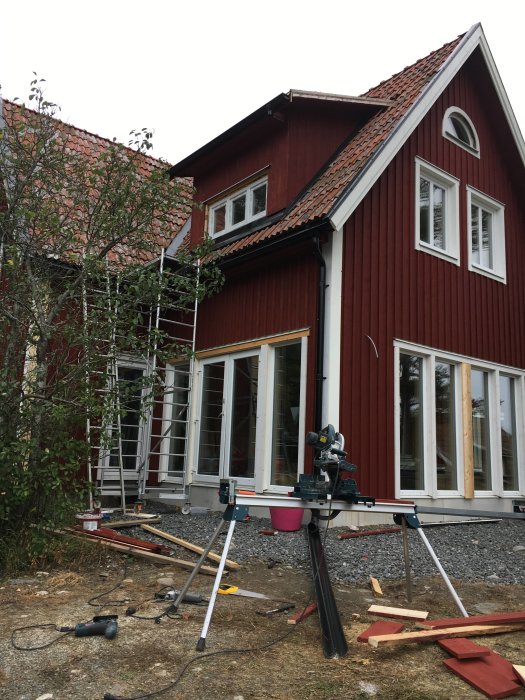 Röd trävilla under renovering med en färdig takkupa, verktyg och byggmaterial på marken.