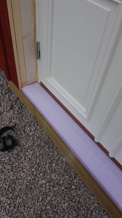 Nyinstallerad bärlina under dörrtröskel med inlagd xps-cellplast för isolering, dörrkarm och grus ute.