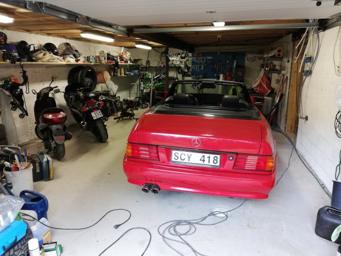 Röd cabrioletbil parkerad i en rörig garageverkstad med motorcyklar och verktyg.