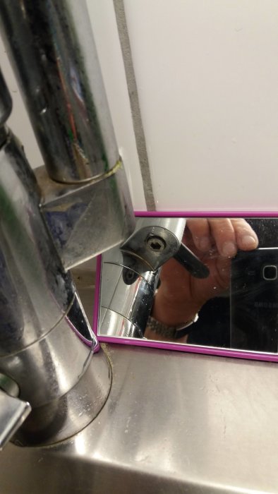 Kökskran med ärgad strålsamlare och synlig insexskruv, spegelreflektion av hand och mobiltelefon.
