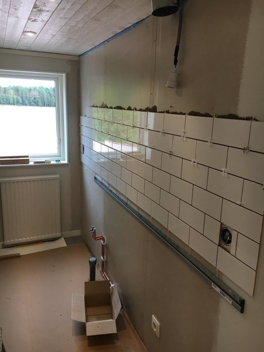 Kök under renovering med nytt kakel som delvis är uppsatt på väggen, rördragning synlig och verktyg på golvet.