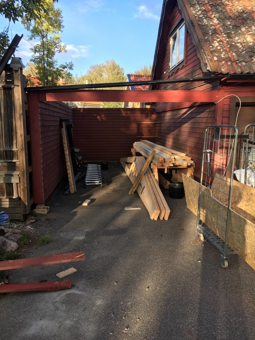 Upprivet carporttak med träbjälkar och byggmaterial utspritt på en uppfart vid en röd trävägg.