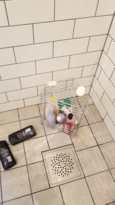Trådkorg med flaskor och behållare på badrumsgolv, två flaskor ligger bredvid.