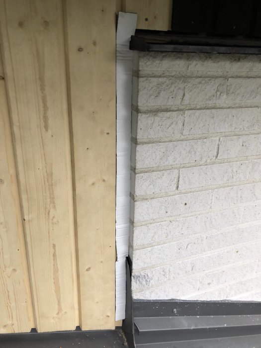 Vit mall av kartong anpassad för montering av träpanel vid husets hörn med bakgrund av tegel och trä.
