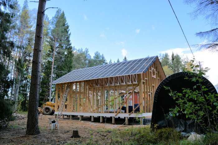 Nybyggt trästomme hus med nylagd taktäckning och byggnadsmaterial i skogsmiljö.