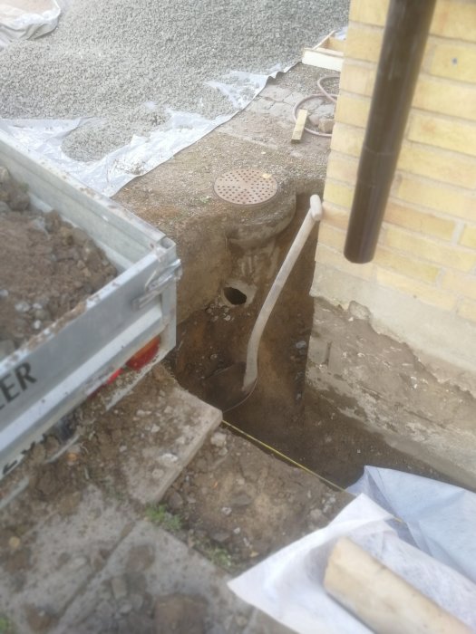 Dräneringsarbete vid brunnen med synlig rörgenomföring och ett överlastat släp med makadam i förgrunden.