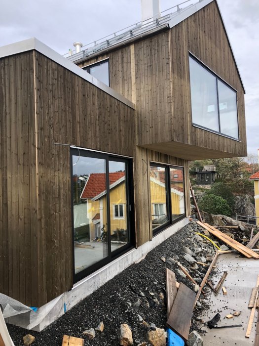 Träfasad på hus med nedmonterad ställning och ojämnt grånad yta, stora fönster, byggmaterial på marken.