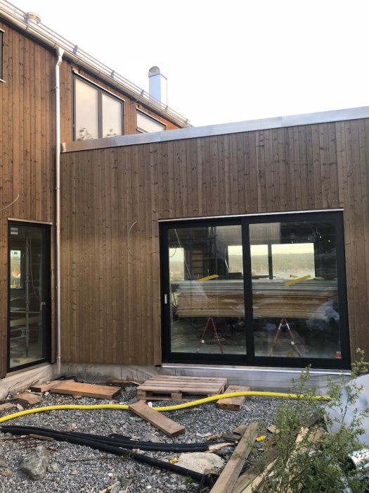 Nedmonterad byggställning vid hus med träfasad och stora fönster, byggmaterial på marken.