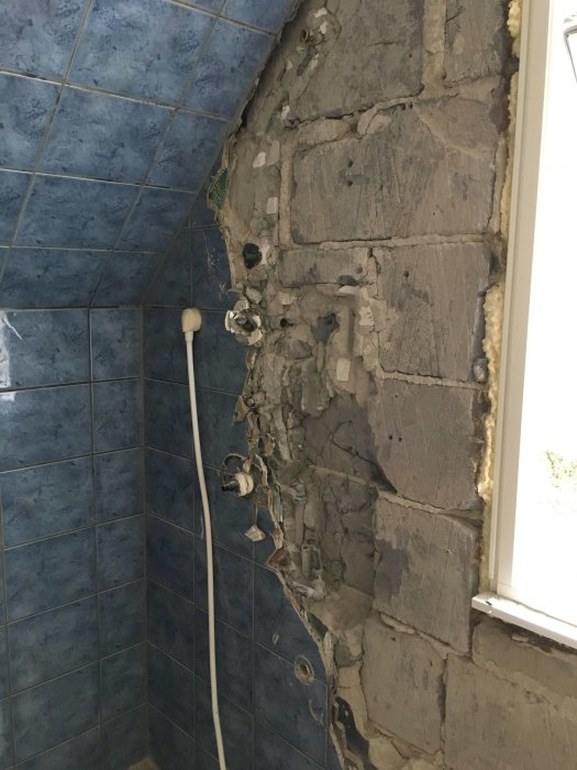 Skadade stenar och sprucket fogmaterial i ett duschutrymme med blå kakel och synliga rörinstallationer.