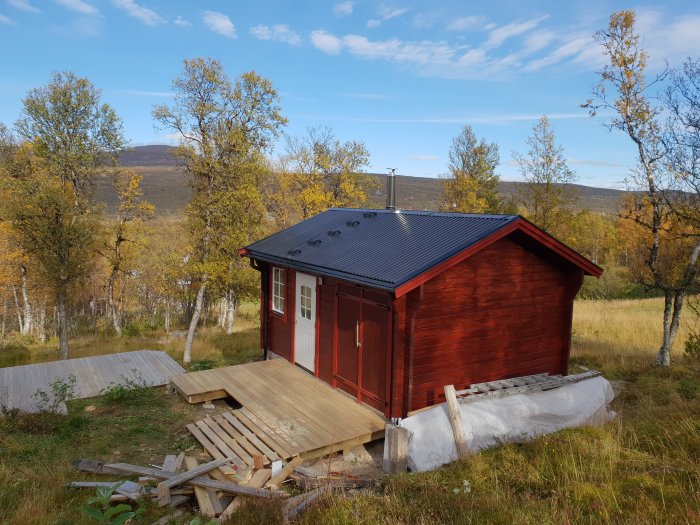 Nymålad stuga med röda väggar och svart tak, omgiven av natur med nybyggt trädäck och material.