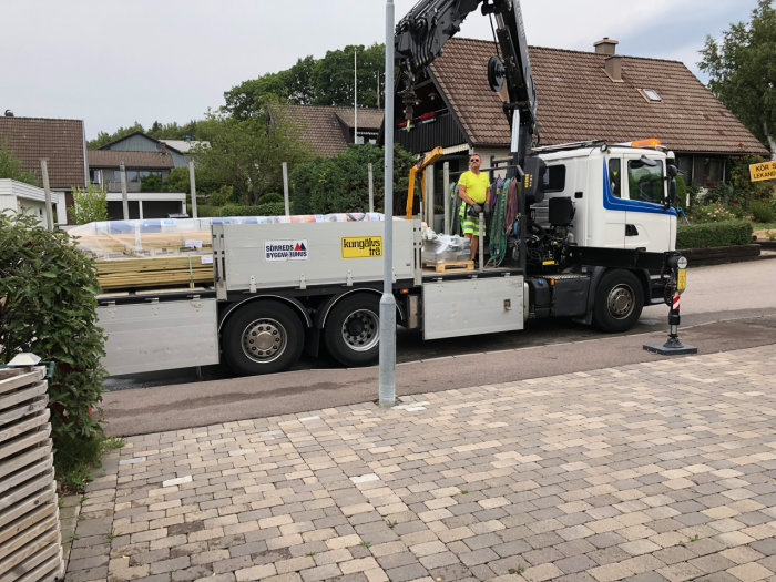 Byggmaterial levereras av en lastbil med kran, förar och hjälpmedel synliga, i ett bostadsområde.