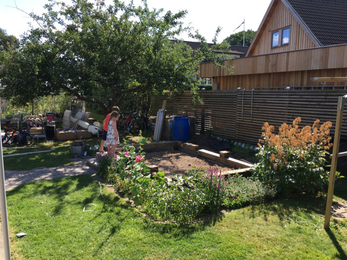 Trädgård med pågående bygge av uteplats; person vattnar blommor vid solig gräsmatta med material och stolpar uppställda.