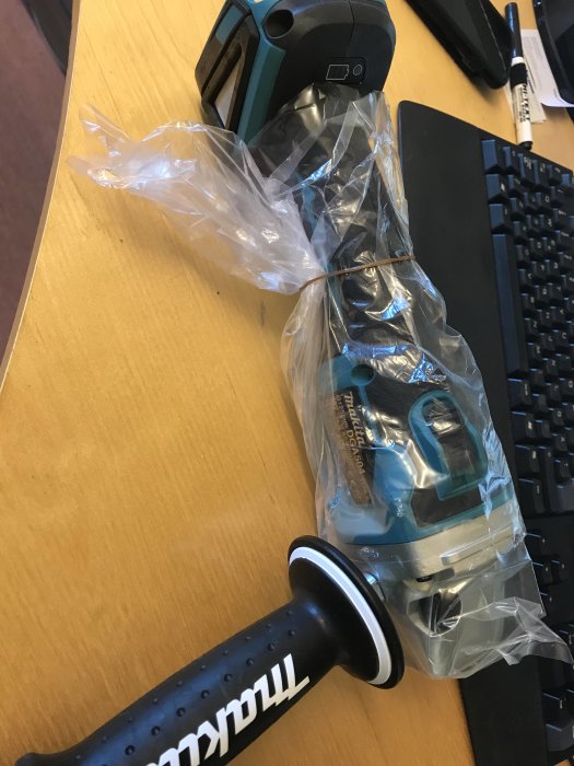 Makita DGA504 sladdlös vinkelslipmaskin delvis i plastförpackning på ett skrivbord med datorutrustning i bakgrunden.