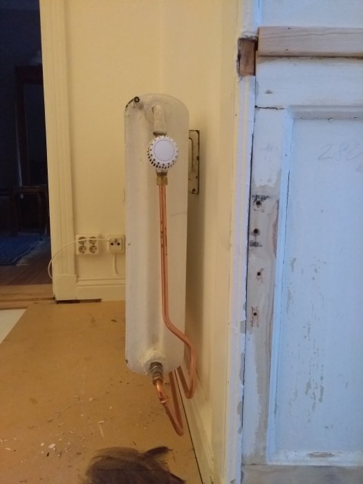 Osäkert upphängt vit element med tunna kopparledningar vid en sliten vit dörrpost i ett renoveringsobjekt.