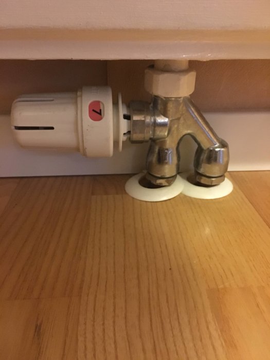 Radiator med fastsittande vit termostat ovanför trägolv och under vit bräda.