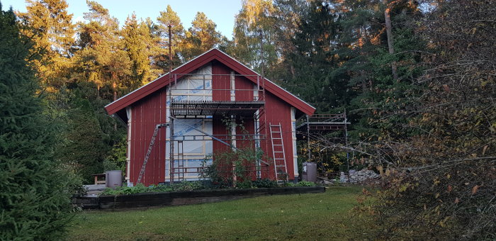 Röd stuga under renovering med ny panel på en gavel och presenningar, omgiven av grönska.