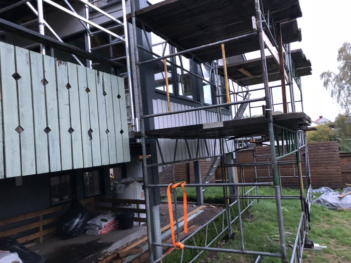 Byggnadsställning utanför ett hus med nyligen uppspikad panel under fönstren i skymningen.