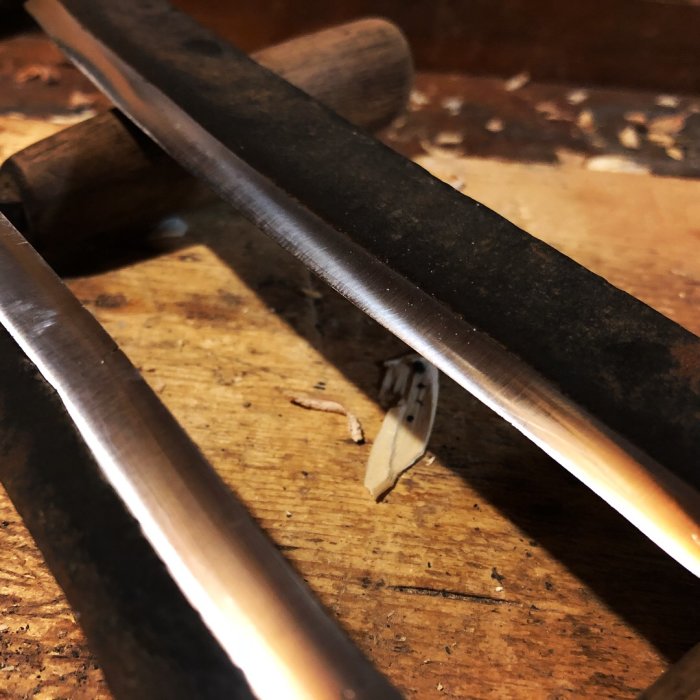 Närbild av två smidda metallverktyg, troligen knivblad, på en träbänk.