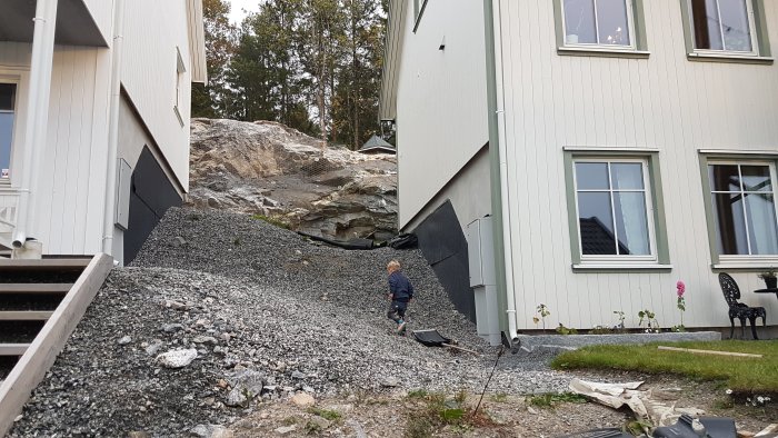 Plats för byggprojekt mellan två hus med grov grus och stenmassor, barn och skottkärra syns.