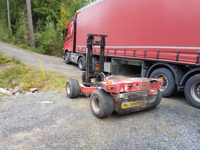 Lastbil med röd släpvagn och en liten gaffeltruck vid sidan, på en grusväg nära skog.