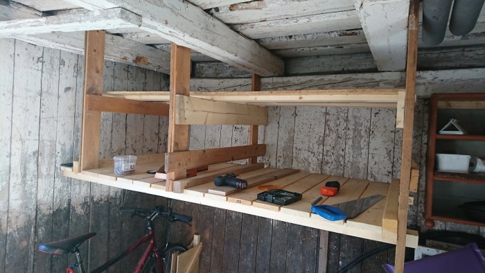 Hyllsystem under konstruktion i lada med verktyg och cykelställ i förgrunden.