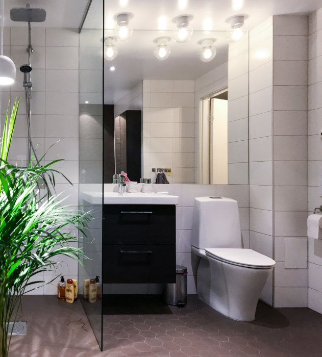 Modernt badrum med spotlights, spegel med armaturer, hängande växt och pendellampa.