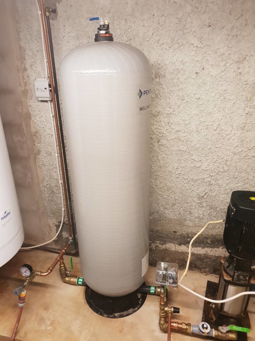 Glasfiberhydrofor med kopplingsrör, manometer och elektrisk pump i källare.