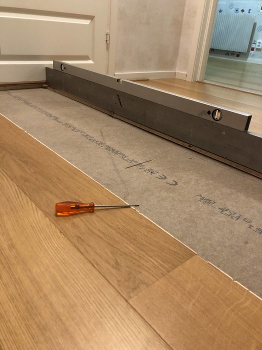 Uppmätt undergolv i hall med låg nivå intill rät skiva och skruvmejsel på trägolv.