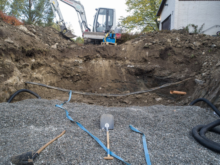 Grävmaskin bredvid en öppen schakt med synlig servisledningskabel och grus på en byggtomt.