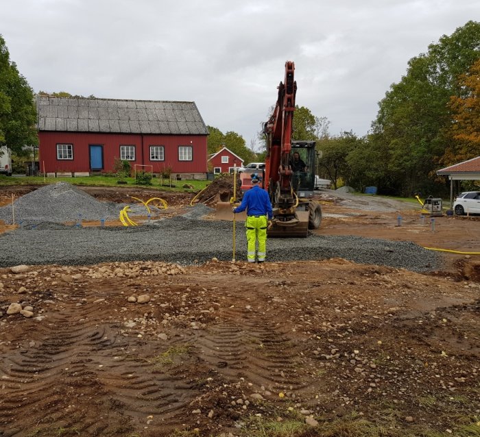 Arbetare skottar makadam vid byggarbetsplats med grävmaskin och skyddsrör i marken.