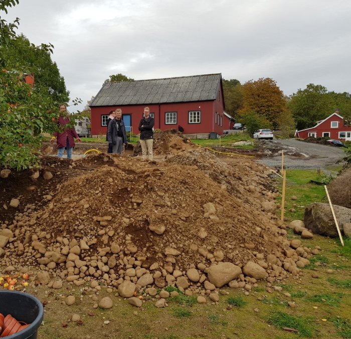 Personer diskuterar vid en hög utskufflad makadam på en byggarbetsplats med röd husbakgrund.