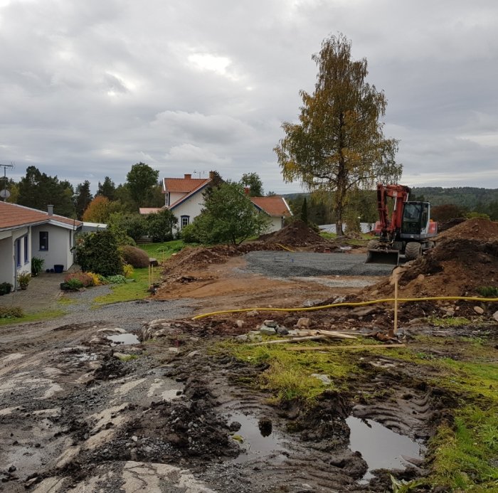 Byggarbetsplats med grävmaskin och högar av makadam, planering av slänt och gångväg pågår.