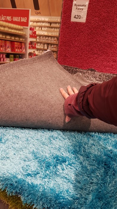 Person som känner på en fluffig, turkos matta bredvid en större grå prydnadskudde och röda mattor i bakgrunden.
