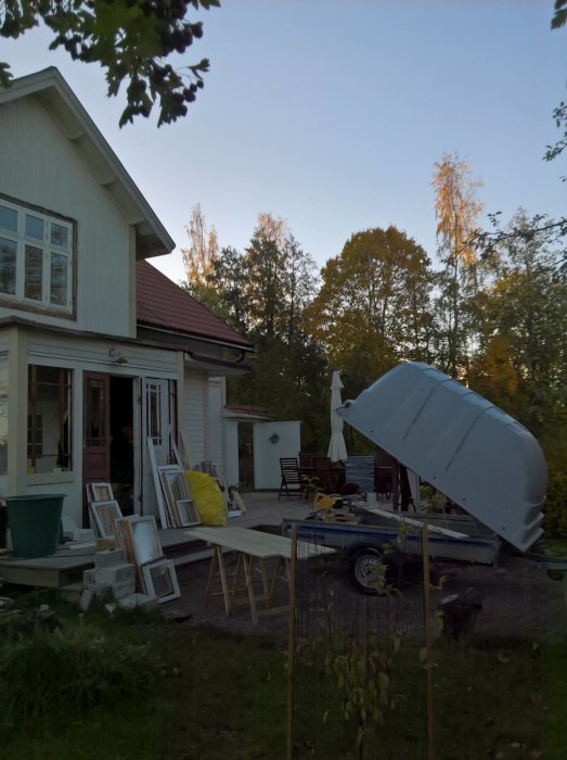 Trädgård med byggmaterial och verktyg utspridda, vit villa i bakgrunden, och en släpvagn i förgrunden.